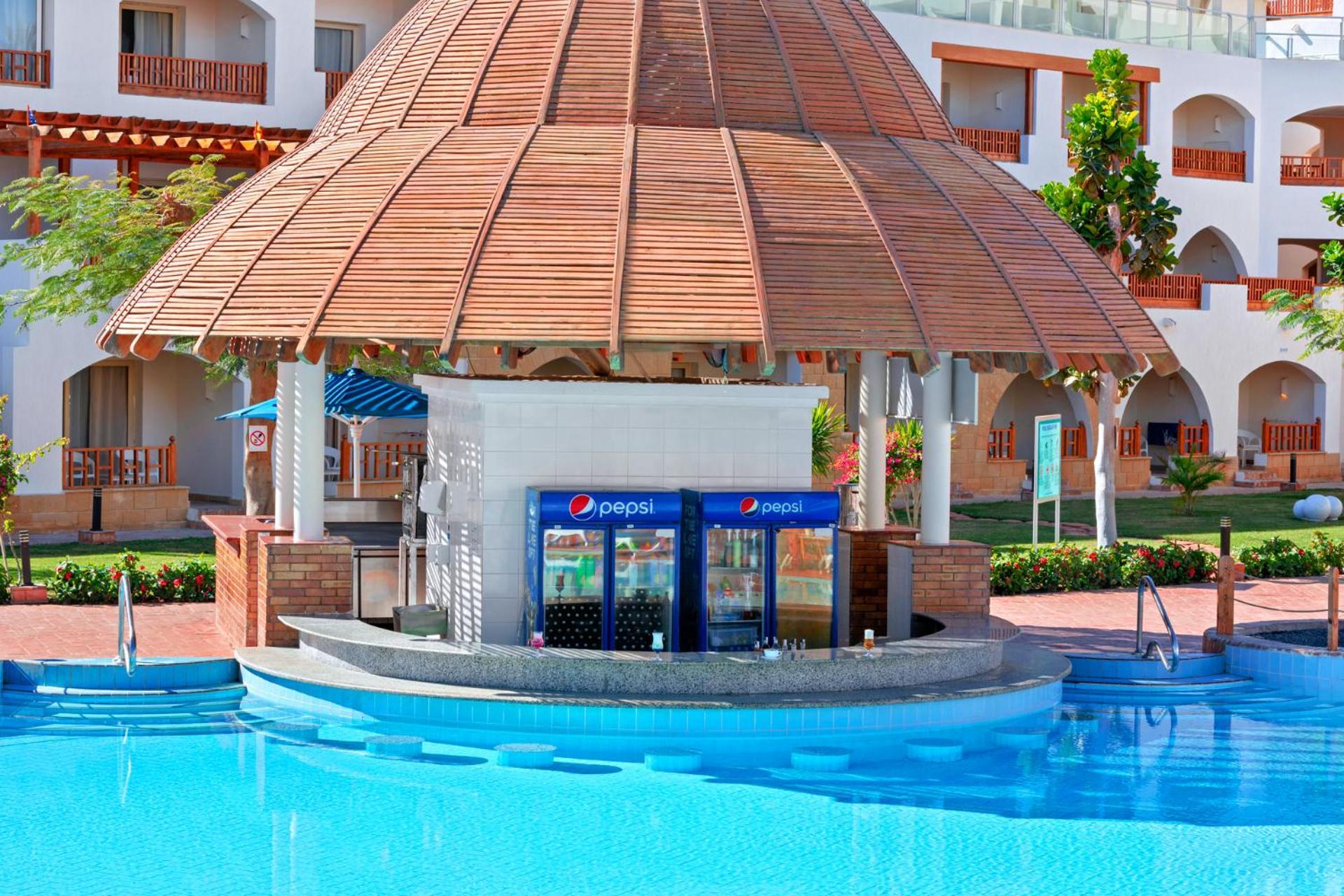 فندق رويال جراند شرم ريزورت شرم الشيخ المظهر الخارجي الصورة The photo shows a poolside bar with a thatched roof structure, located near a swimming pool. The bar features a counter where drinks are served, and there are vending machines displaying the Pepsi brand. Surrounding the bar, there are neatly landscap